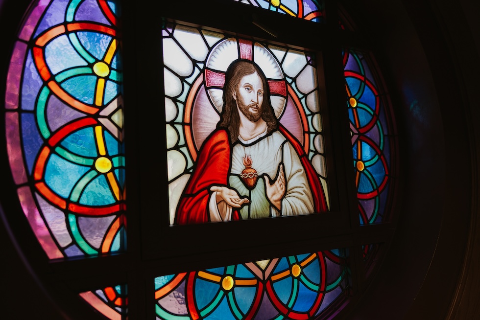 Image of a stained glass with Jesus in front of a cross and his hand held out in front.